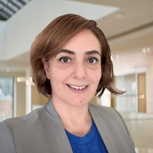 BMS employee Ajlan Atasoy, MD has short brownish, red hair and wears a grey blazer over a blue blouse. She is in an office setting.